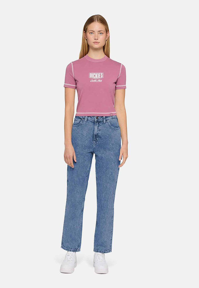 Jovem mulher em pé, vestindo uma blusa cropped rosa da Dickies, calças de ganga de cintura alta azuis e ténis brancos, contra um fundo claro e liso.