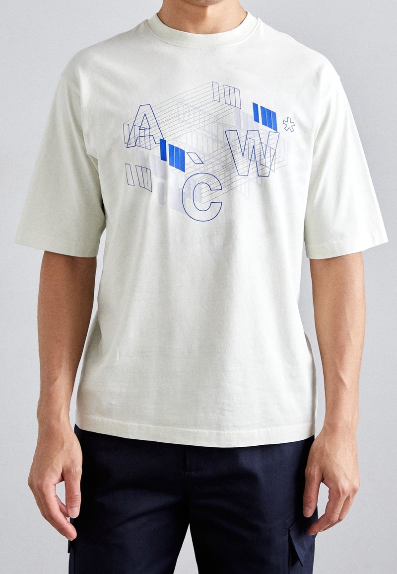 Muž v bílém tričku s krátkým rukávem, na kterém je modrošedý abstraktní geometrický design a písmena "A", "C", "W" na přední straně.