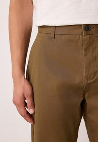 Bruine katoenen broeken met een gladde textuur, voorzien van een knoopsluiting en zijzakken. Het ontwerp bevat subtiele stikseldetails.