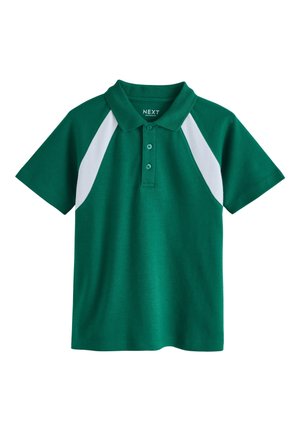 Blonder Junge steht vor Holzwänden, trägt ein grünes Poloshirt mit weißen Akzenten, hellblaue Jeans und weiße Turnschuhe.