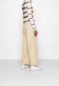 Pantalones beige de pierna ancha de algodón, combinados con una camiseta de manga larga con efecto tie-dye en crema y negro. Zapatillas deportivas blancas de caña alta completan el conjunto.