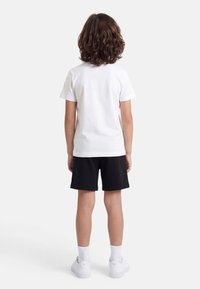 Παιδί με ώμος μήκος σγουρά μαλλιά, που φοράει λευκό μπλουζάκι, μαύρο σορτς, λευκές κάλτσες και λευκά αθλητικά παπούτσια, στέκεται γυρισμένο με την πλάτη σε λευκό φόντο.