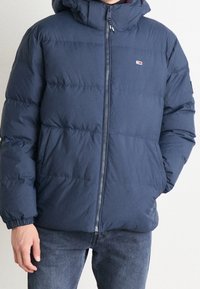 Mörkblå pufferjacka med dragkedja fram, huva, elastiska ärmslut och kontrasterande logotyp. Har kviltad textur och sidofickor.