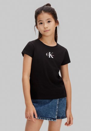 Jong meisje met donker haar, gekleed in een zwart Calvin Klein T-shirt en een blauwe denimrok, staand tegen een effen grijze achtergrond.