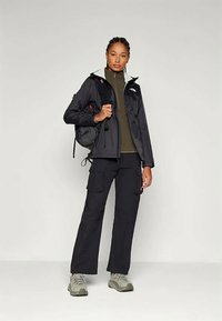 Melnā ūdensizturīgā jaka ar pelēku kapuci, olīvu zaļš iekšējais slānis, melni šortai un pelēki pārgājienu apavi; redzams ar mazu melnu mugursomu.