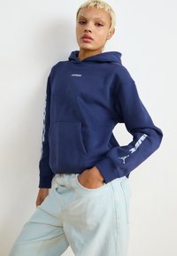 Hoodie azul-marinho com bolso frontal, apresenta um logotipo branco no peito e na manga, feito de tecido macio, combinado com jeans azul-claro.