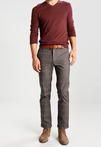 Bordowy sweter z dekoltem w serek, szare spodnie w kratę z czerwonymi i czarnymi akcentami, brązowy skórzany pasek oraz jasnobrązowe botki za kostkę.