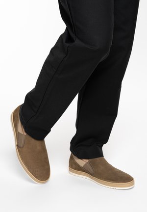 Person trägt schwarze Hose und braune lässige Slipper mit weißen Sohlen, steht mit einem leicht angehobenen Fuß.