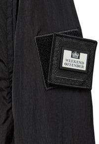 Black fabric coat detail featuring a prominent embroidered patch with "WEEKEND OFFENDER" in white text, framed by a textured black border.