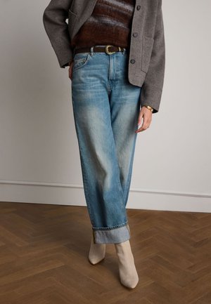 Jeans en denim bleu délavé avec une coupe décontractée, ourlets retroussés et une légère brillance. Associés à des bottines à talons beige clair. Veste grise texturée.