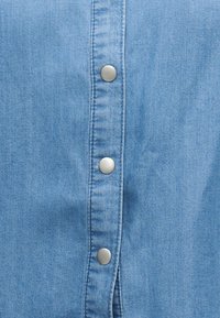 Lichtblauw denimstof met een verticale rij van drie zilveren drukknopen, met zichtbare textuur en stikdetails.