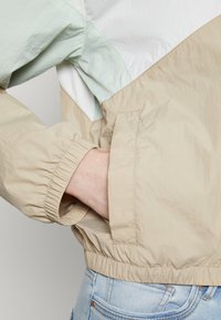 Lekka kurtka w odcieniach beżu i jasnoniebieskim. Elastyczne mankiety i dół. Posiada kieszeń z przodu. Tekstura wydaje się gładka z lekkim połyskiem.