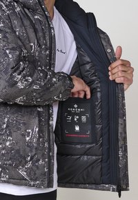 Chaqueta de plumas con un patrón de camuflaje en gris y negro, que presenta un suave forro interior y una etiqueta en el bolsillo interno.