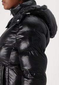 Black padded jacket featuring a glossy finish, high collar, adjustable hood, and a discreet logo patch on the left shoulder.