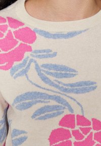 Gestrickter Pullover mit cremefarbenem Grundton, großen rosa Blumenmustern und blauen Blättern, der eine weiche Textur und einen runden Ausschnitt aufweist.