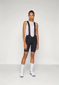 Μαύρες ποδηλατικές φόρμες με ρυθμιζόμενες τιράντες, με αντίθετο γκρίζο λογότυπο. Φορεμένες πάνω από ένα ανοιχτόχρωμο αμάνικο τοπ. Λείο ύφασμα.