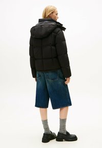Zwarte pufferjack met een capuchon, doorgestikte ontwerp en elastische manchetten, gecombineerd met blauwe denimshorts. Het model draagt grijze sokken en zwarte instappers.