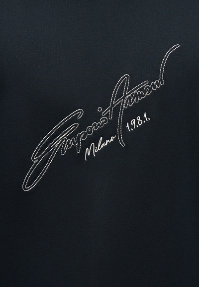 Λευκή ραφή σε στυλ γραφής πάνω σε μαύρο ύφασμα γράφει "Giorgio Armani Milano 1.9.8.1."