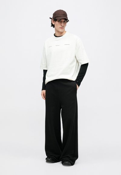 Biela nadrozmerná tričko s jemným nápisom, vrstvené cez čierne tričko s dlhým rukávom. Voľné čierne nohavice a tmavohnedá čiapka dopĺňajú vzhľad.