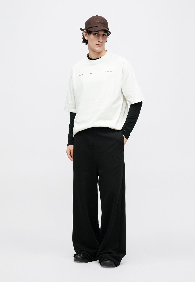 Maglietta bianca oversize con testo sottile, indossata sopra una maglia a maniche lunghe nera. Pantaloni neri larghi e un berretto marrone scuro completano il look.