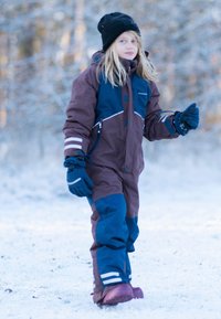 Brun och blå vinteroverall med vita accenter, huva och reflexstripes; kombinerad med en svart mössa och blåa handskar; snöig bakgrund.