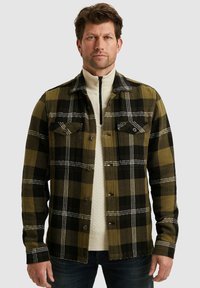 Geblokte shirt in groen, zwart en wit met een visgraatpatroon, voorzien van twee borstzakken en knopen, gedragen over een crèmekleurige trui.