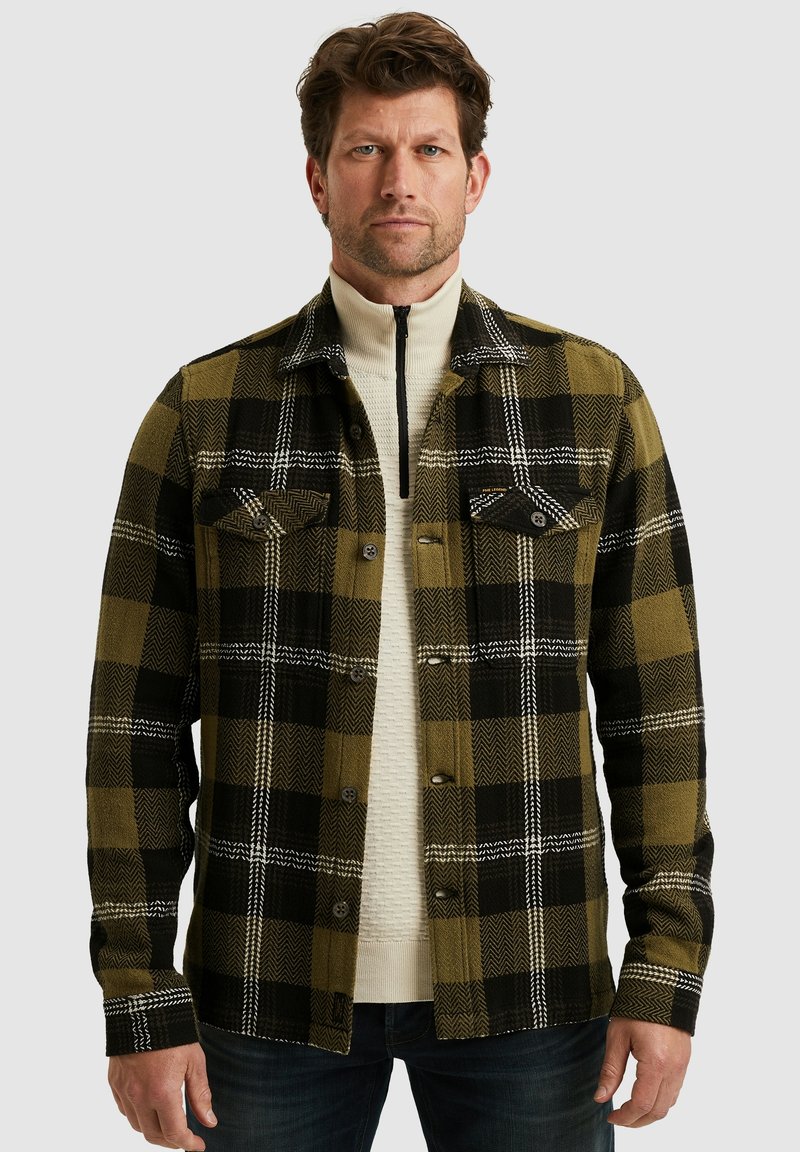 Geblokte shirt in groen, zwart en wit met een visgraatpatroon, voorzien van twee borstzakken en knopen, gedragen over een crèmekleurige trui.