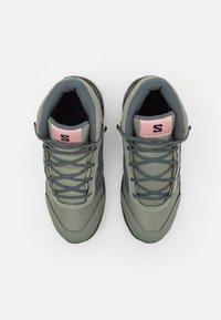 Zapatillas verdes con acabado texturizado, paneles de malla y cordones grises. Detalle rosa en el talón; presenta el logo en la lengüeta.