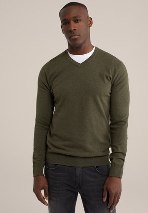 Grüner V-Ausschnitt-Pullover aus strukturiertem Strick, mit langen Ärmeln sowie gerippten Bündchen und Saum, getragen über einem weißen T-Shirt, kombiniert mit dunklen Jeans.