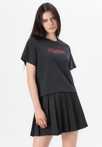 Black short-sleeve t-shirt with a red Coca-Cola logo printed on the front, paired with a pleated black skirt, showcasing a relaxed fit.