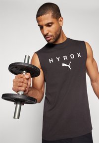 Black sleeveless athletic shirt with white "HYROX" text and Puma logo. Person holding a black weight with a metal handle.