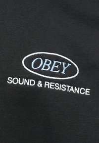 Črn material z vezenim napisom "OBEY" v svetlo modri barvi in "SOUND & RESISTANCE" v beli, s gladko teksturo in ovalnim oblikovanjem.
