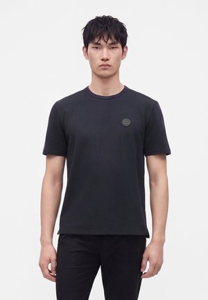 Junger Mann trägt ein einfarbiges schwarzes T-Shirt mit kurzen Ärmeln und kleinem rundem Logo auf der linken Brust, steht vor einem schlichten weißen Hintergrund.
