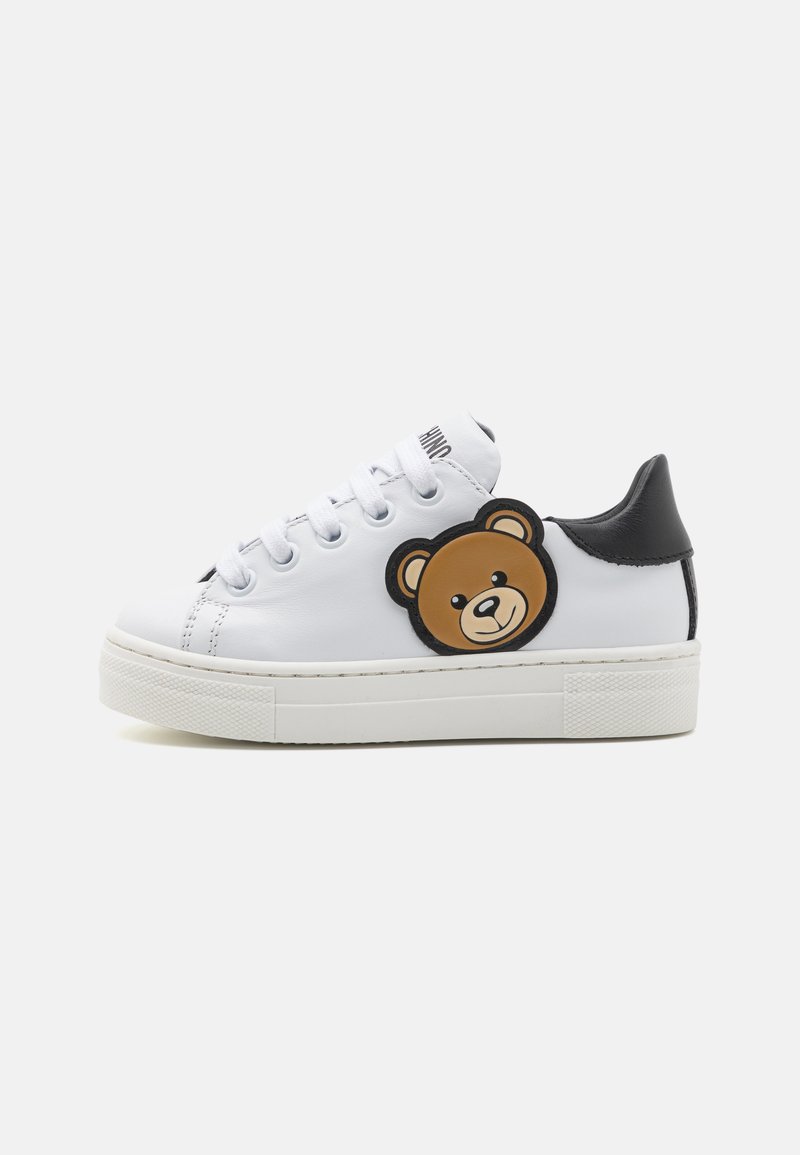 Baskets blanches avec talon noir, arborant un patch en forme de tête d'ours rond sur le côté, semelle texturée et design à lacets.