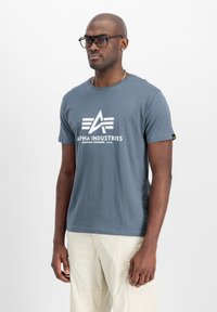 Graues Baumwoll-T-Shirt mit einem weißen Grafiklogo "ALPHA INDUSTRIES" auf der Vorderseite, kurzen Ärmeln und einem Rundhalsausschnitt mit flachem Saum.