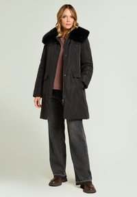 Chaqueta acolchada negra con cuello de piel, cierre de cremallera, dos bolsillos delanteros y corte recto. Llevada sobre una camiseta marrón y vaqueros grises.