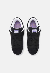 Baskets en daim noir avec semelles blanches. Elles présentent des lacets proéminents, une languette brandée violet et blanc, et un motif de semelle intérieure texturé.