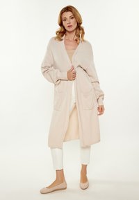 Cardigan lungo aperto in morbido tessuto beige a maglia, con due tasche frontali, polsini a coste e una silhouette rilassata.