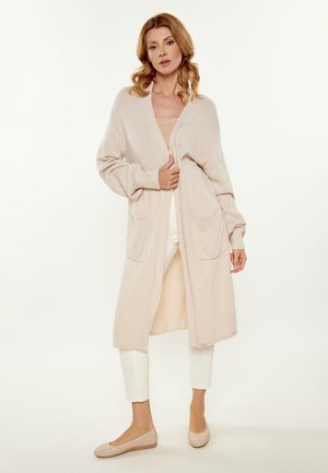 Cardigan lungo aperto in morbido tessuto beige a maglia, con due tasche frontali, polsini a coste e una silhouette rilassata.