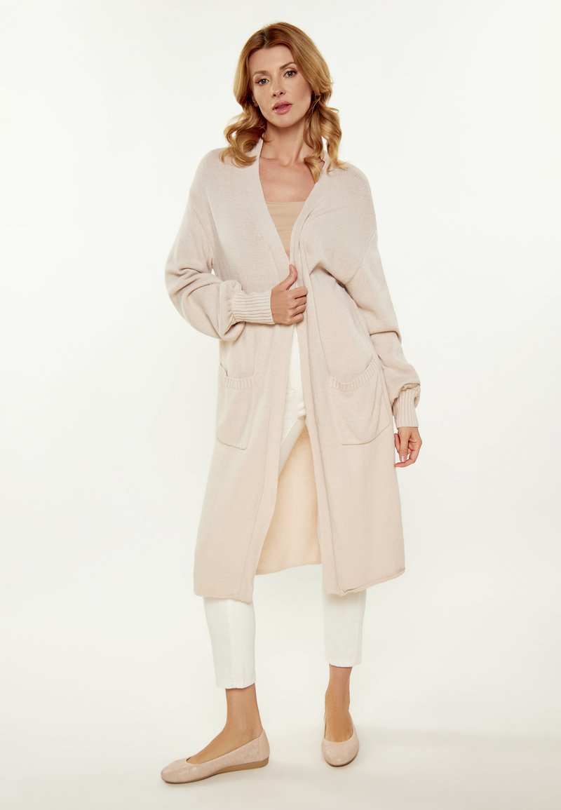 Cardigan lungo aperto in morbido tessuto beige a maglia, con due tasche frontali, polsini a coste e una silhouette rilassata.