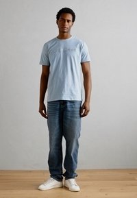 Lichtblauw t-shirt met geborduurde tekst, gecombineerd met vervaagde blauwe jeans en witte sneakers. Model staat tegen een effen achtergrond.