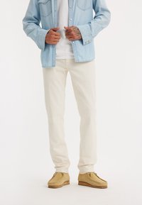 Lichtdenim overhemd, witte T-shirt, crèmekleurige broek en tan suedineschoenen. Heeft twee borstzakken en lange mouwen opgerold.
