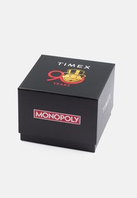 Melna kaste ar drukātu dizainu. Satur "TIMEX" un "90 GADI" baltā un sarkanā krāsā, kā arī dzeltena Monopola varoņa ilustrācija. Sarkanā "MONOPOLY" logo.