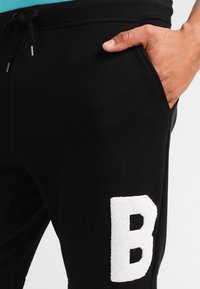 Pantalons de survêtement noirs avec une lettre 'B' texturée blanche sur la jambe droite, avec une taille élastique et une poche latérale visible.