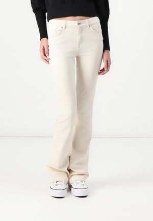 Femme portant un jean bootcut écru, un pull court noir et des baskets blanches, debout devant un fond blanc uni.