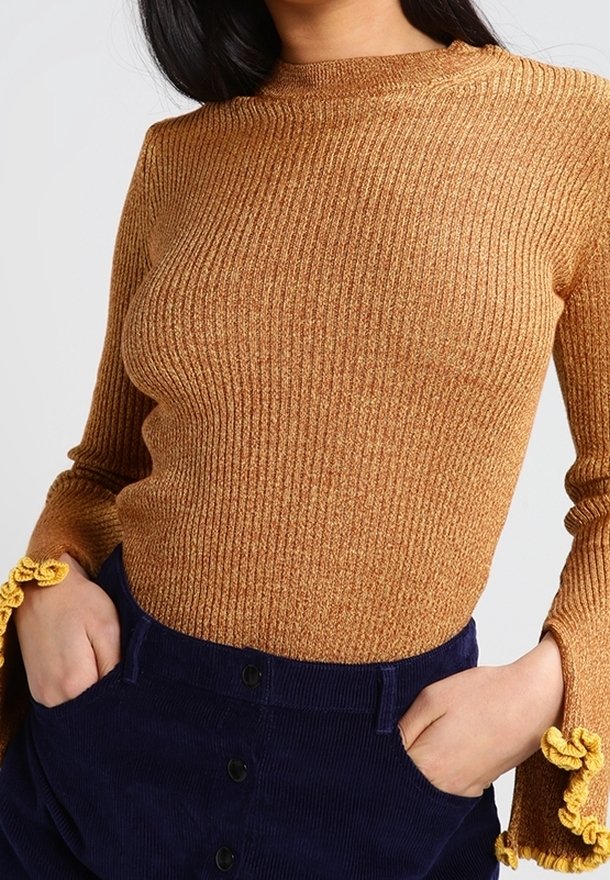 Sweter o prążkowanej fakturze w złocistobrązowym kolorze, z wysokim kołnierzem i rozkloszowanymi rękawami z żółtymi akcentami szydełkowymi; zestawiony z granatową spódnicą sztruksową.