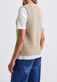 Beżowy dziergany top bez rękawów noszony na białej koszulce, z prążkowaną teksturą i luźnym krojem, zestawiony z niebieskimi spodniami dżinsowymi.