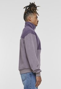 Sudadera de forro polar bicolor morada con cuello alto y cierre de cremallera, que presenta una textura suave y puños acanalados, combinada con jeans de mezclilla azules.