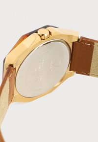 Orologio dorato con cassa ottagonale, cinturino in pelle marrone e fondo in acciaio inossidabile rotondo. Presenta una corona texturizzata e dettagli incisi.