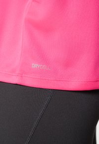 Roosa sportlik särk, mis on tehtud siledast kangast ja millel on ääre lähedal "DRYCELL" logo, kombineeritud tekstureeritud mustade retuusidega.